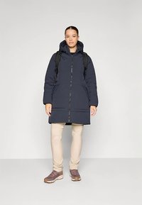 Paminkštintas tamsiai mėlynas paltas iki kelių su kapišonu, užtrauktuku priekyje ir dviem šoninėmis kišenėmis. Dėvimas ant šviesiai smėlinio aprangos ir pilkai rudi bateliai.