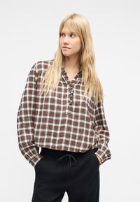 Ξανθιά γυναίκα που φοράει κόκκινο και λευκό πουκάμισο με μακριά μανίκια καρό και μαύρο παντελόνι με κορδόνι, με τα χέρια στις τσέπες, κοιτάζοντας στο πλάι.