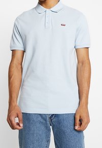 Ljusblå polo-tröja i texturerad bomull, med en tre-knappsslutning och en liten röd logotyp på vänster bröst.