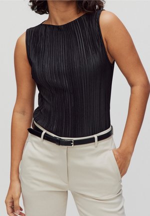 Femme portant un haut noir sans manches plissé rentré dans un pantalon beige taille haute avec une ceinture noire, une main dans la poche, sur un fond uni.