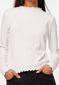 Vit ribbad långärmad topp med en scallop-formad halsringning och nederkant. Materialet har en mjuk textur; åtsittande design med långärmar och sidofickor.