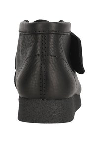 Svart läderankleboot med en texturerad yta, matt svart gummisula och en platt bakdel. Har en kontrasterande svart mockaaccent.