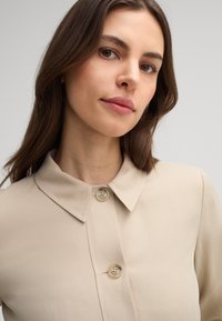 Camicia beige con colletto e chiusura a bottone, caratterizzata da una texture di tessuto liscia e dettagli di cucitura sottili.