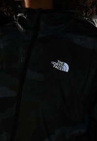 Chaqueta de camuflaje negra con cremallera frontal, capucha ajustable y el logo de The North Face en blanco en la parte izquierda del pecho.