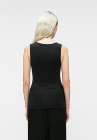 Oseba z dolgimi svetlimi lasmi, oblečena v brezrokavnik črne barve in črne hlače, obrnjena stran od fotografa pred enostavno belo ozadjem.