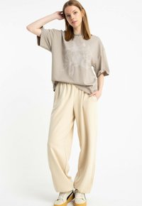 Młoda kobieta stoi z ręką na głowie, ubrana w beżowy t-shirt z nadrukiem smoka, kremowe szerokie spodnie i białe trampki z gumowymi podeszwami.