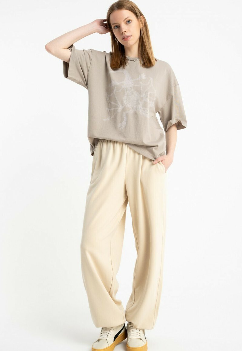 Młoda kobieta stoi z ręką na głowie, ubrana w beżowy t-shirt z nadrukiem smoka, kremowe szerokie spodnie i białe trampki z gumowymi podeszwami.