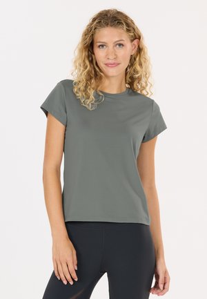 Vrouw met krullend blond haar, gekleed in een grijs T-shirt met korte mouwen en zwarte leggings, staand tegen een effen witte achtergrond.