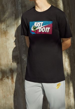 Osoba w czarnej koszulce z kolorowym napisem „JUST DO IT” i kreskówkową grafiką, w białych spodniach z żółtym logo Nike.