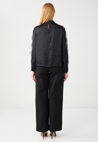 Schwarze Bluse mit Spitzendetails an den Ärmeln, mit einem Schlitz und Knopfverschluss auf der Rückseite, kombiniert mit schwarzen Hosen mit weitem Bein.