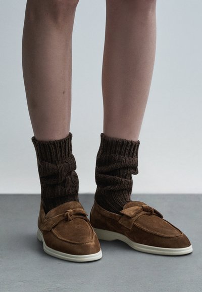 Smeđe antilop mokasinke s detaljem u obliku mašne, uparene s debelim, teksturiranim smeđim pletenim čarapama koje imaju rebrasti uzorak oko lista.