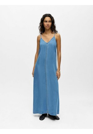Femme portant une robe longue en denim bleu clair sans manches avec de fines bretelles et des chaussures noires, debout sur un fond blanc uni.