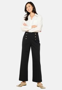 Črne hlače širokega kroja z zlatimi gumbnimi detajli, v kombinaciji z belo srajco z dolgimi rokavi. Obleko dopolnjujejo zlate ravne čevlje.