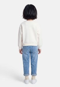 Enfant aux cheveux noirs courts, portant un sweat-shirt blanc, un jean bleu clair retroussé, des chaussettes beiges et des baskets blanches, debout de dos.