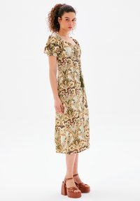 Blumenmuster-Midi-Kleid mit Puffärmeln, in Grün- und Brauntönen, quadratischem Ausschnitt und fließender Silhouette. Kombiniert mit braunen Plateau-Sandalen.