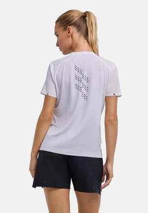 Femme blonde avec une queue de cheval tournée de dos, portant un t-shirt de sport blanc avec des fentes de ventilation noires et un short noir, sur fond uni.