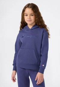 Ragazzina con capelli ricci, che indossa un hoodie blu Champion e pantaloni della tuta coordinati, in piedi davanti a uno sfondo bianco.