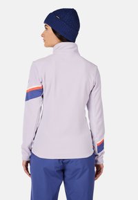 Giacca in pile viola chiaro con colletto alto, caratterizzata da strisce blu e arancioni sulle maniche. Indossata con un berretto lavorato a maglia blu scuro.
