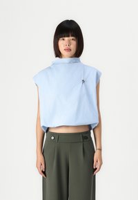 Cropped top azzurro chiaro con colletto alto e senza maniche, realizzato in tessuto liscio, presenta una spilla decorativa. Abbinato a pantaloni verdi a vita alta.