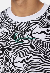 Bílé tričko s černým vířivým vzorem, kulatým výstřihem a zeleným vyšívaným detailem postavy a textu.