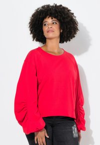 Rotes Sweatshirt mit langen Ärmeln und gerafften Ballonärmeln, rundem Halsausschnitt und cropped Bund. Kombiniert mit dunklen Denim-Jeans.