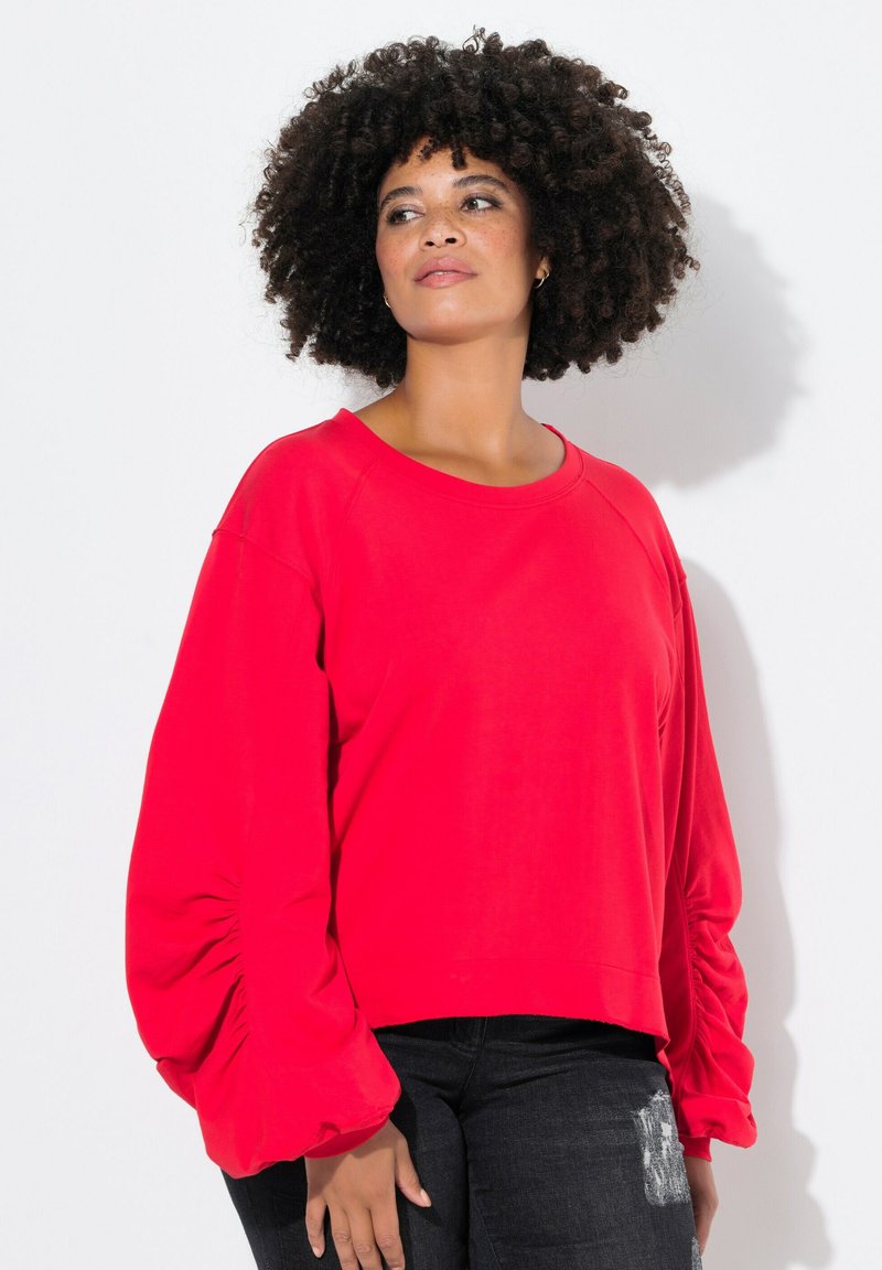 Rotes Sweatshirt mit langen Ärmeln und gerafften Ballonärmeln, rundem Halsausschnitt und cropped Bund. Kombiniert mit dunklen Denim-Jeans.