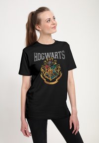 Czarny t-shirt z kolorowym herbem Hogwartu, z złotymi akcentami i napisem "HOGWARTS" wytłuszczonym szarym drukiem nad herbem.