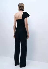 Black jumpsuit with a one-shoulder design and pleated detail. Features a fitted bodice and wide-leg pants, smooth fabric texture.