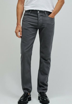 Hombre con pantalones rectos gris oscuro, camisa blanca y mocasines de cuero negros, de pie con una mano en el bolsillo.