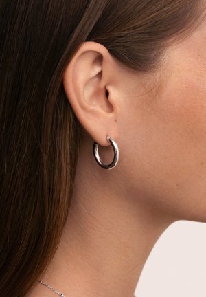Boucle d'oreille en métal argenté avec une finition polie. Le design circulaire se fixe solidement sur le lobe, mettant en valeur une surface lisse et réfléchissante.