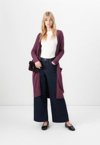 Vrouw in een witte geribbelde top, lange paarse cardigan, wijde donkere spijkerbroek, zwarte flats en met een kleine donkere handtas.