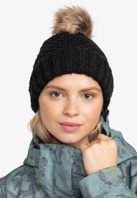 Gorro de punto negro con un diseño de canalé texturizado y un pom-pom de piel sintética en la parte superior, llevado por una persona con una chaqueta de color teal con un patrón.