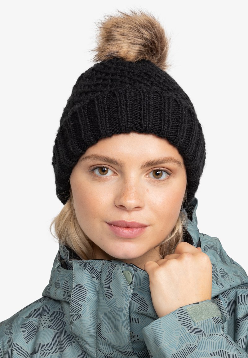 Gorro de punto negro con un diseño de canalé texturizado y un pom-pom de piel sintética en la parte superior, llevado por una persona con una chaqueta de color teal con un patrón.