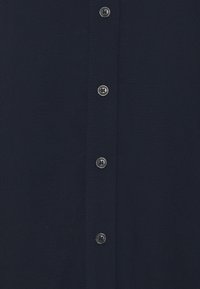 Marineblaue Hemd mit einer glatten Textur, das in der Vorderseite mit einer Reihe von sechs silbernen Knöpfen ausgestattet ist. Der Stoff wirkt leicht und weich.