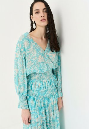 Femme aux cheveux foncés portant une robe longue bleue à fleurs avec taille smockée et volants, debout sur un fond blanc uni.