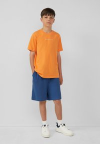 Oranje katoenen T-shirt met korte mouwen en witte tekst, gecombineerd met blauwe katoenen shorts. Model draagt witte sneakers en sokken.