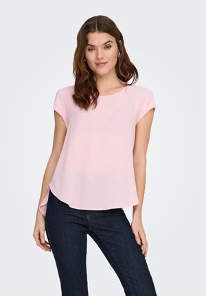 Young woman wearing light pink short-sleeve blouse and dark blue jeans, standing with a slight smile and relaxed posture.