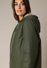 Veste imperméable vert olive avec capuche, dotée d'une fermeture éclair frontale argentée et d'une texture lisse et brillante, adaptée à une tenue décontractée.