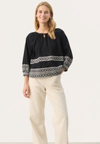 Zwarte katoenen blouse met een ronde halslijn en strikdetail, met witte geborduurde geometrische patronen op de mouwen en zoom. Gepaard met beige broek.
