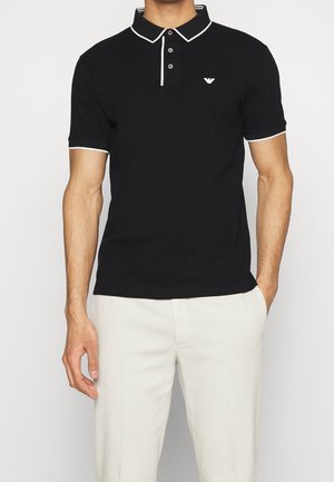 Uomo che indossa una polo nera con bordatura bianca e logo sul petto, abbinata a pantaloni bianchi, con una mano in tasca, in piedi contro uno sfondo semplice.