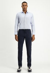 Camicia button-up blu chiaro con un motivo sottile, abbinata a pantaloni con vita elastica blu scuro e sneakers grigie con accenti neri.