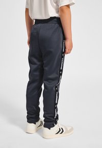 Granatowe spodnie sportowe z elastycznym paskiem w talii, boczne paski z białym logo „hummel”, gładka faktura i zwężany krój nogawek.