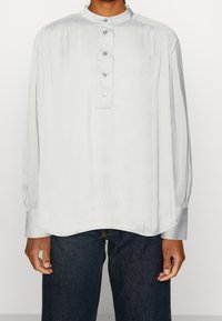 Blusa a maniche lunghe grigia chiara con colletto alla mandarino, chiusura con bottoni e dettagli arricciati sulle spalle e sulle maniche. Texture morbida.