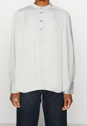 Blouse longue gris clair à manches longues avec un col mandarin, fermeture bouton-pression à l'avant et détails froncés aux épaules et aux manches. Texture douce.