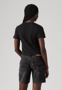 Schwarzes, kurzes T-Shirt mit kurzen Ärmeln, kombiniert mit dunkelgrauen Denim-Shorts. Auffällige Details sind sichtbare Nähte und Gesäßtaschen.