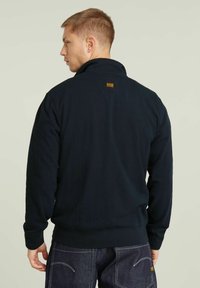 Felpa in pile blu navy con collo alto. Presenta un piccolo logo sul retro e polsini e fondo a costine. Texture liscia, design casual.