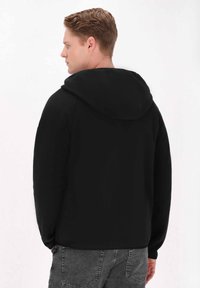 Svart huvtröja med en slät yta, med en stor huva och långa ärmar. Synliga sömmar längs ryggen. Bärs tillsammans med grå jeans.