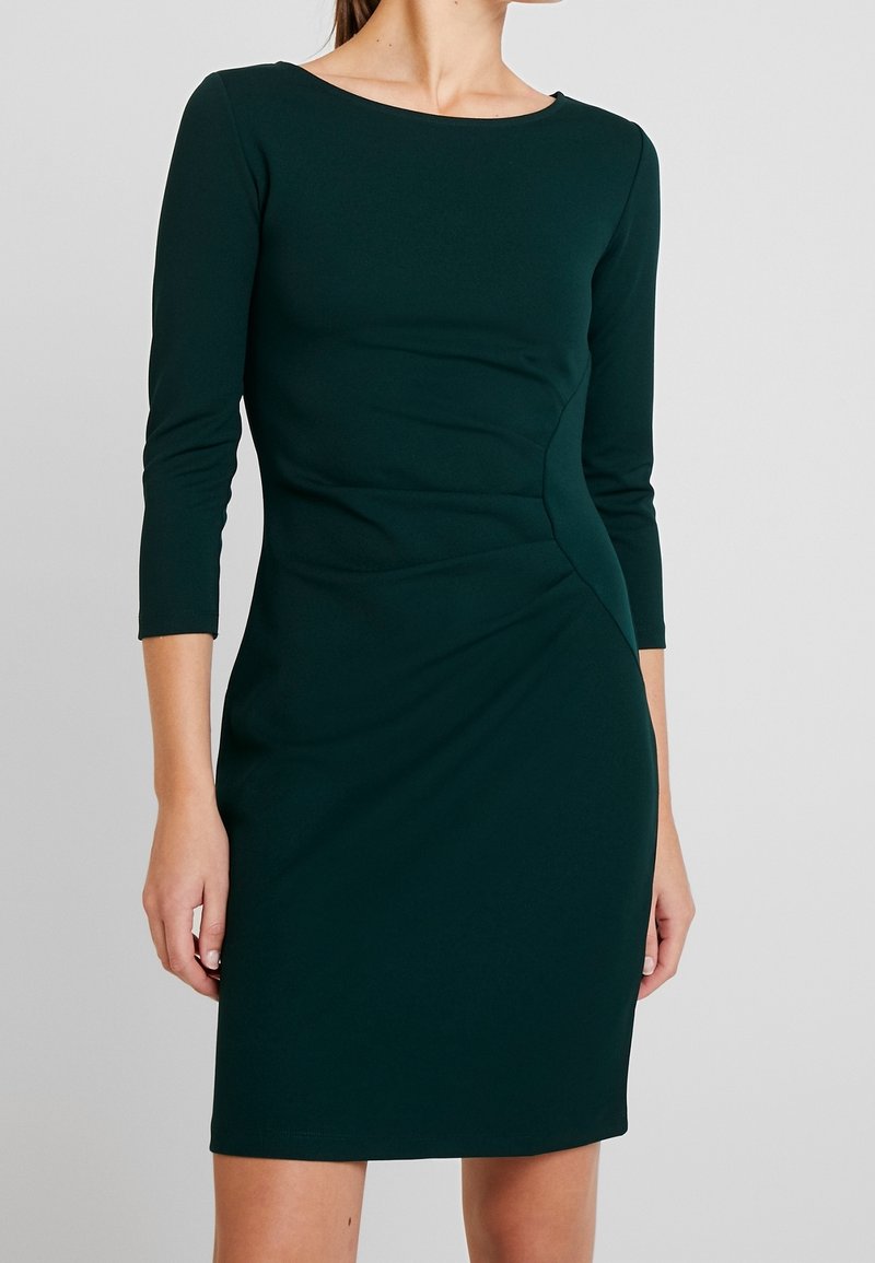 Femme portant une robe ajustée, longueur genou, vert foncé, avec des manches trois-quarts et des détails de couture asymétriques à la taille.