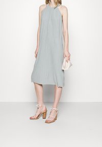 Vestido sin mangas de un gris claro con una tela texturizada, de longitud hasta la mitad de la pantorrilla, combinado con sandalias de tacón blanco con tiras y un bolso pequeño beige.