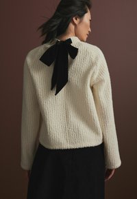 Suéter texturizado color crema con mangas largas y un detalle de lazo de terciopelo negro en la parte posterior. La tela tiene una apariencia suave y acanalada.